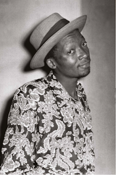 Black and white photo in which a man looks wryly into camera, a hat on his head and a floral button up shirt on.