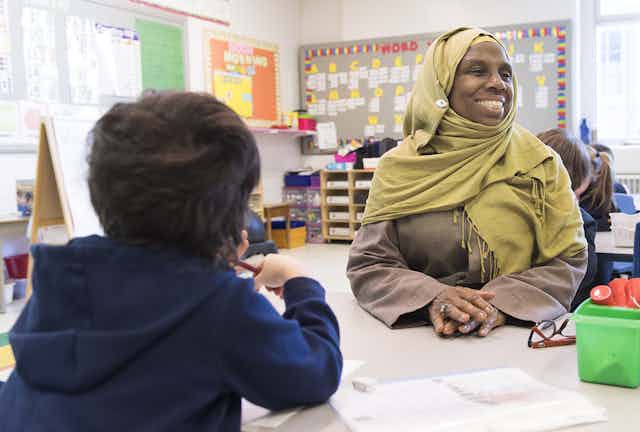 Une femme portant le voile, tout sourire, est assise dans une classe, face à un jeune garçon