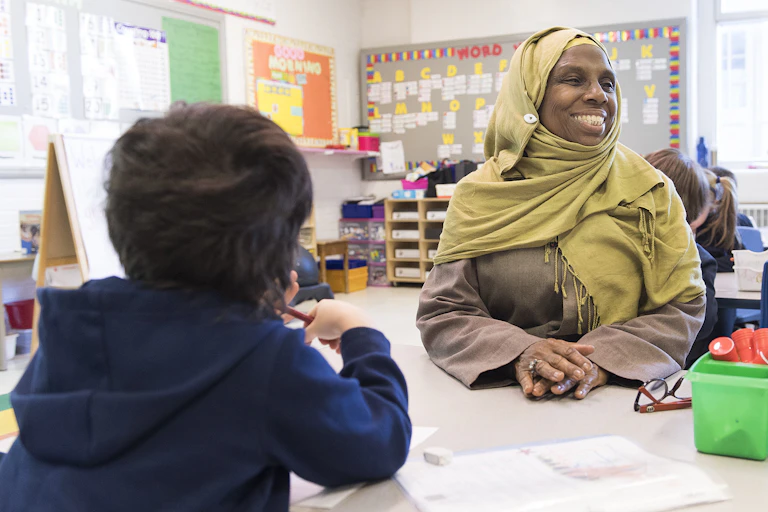 Une femme portant le voile, tout sourire, est assise dans une classe, face à un jeune garçon