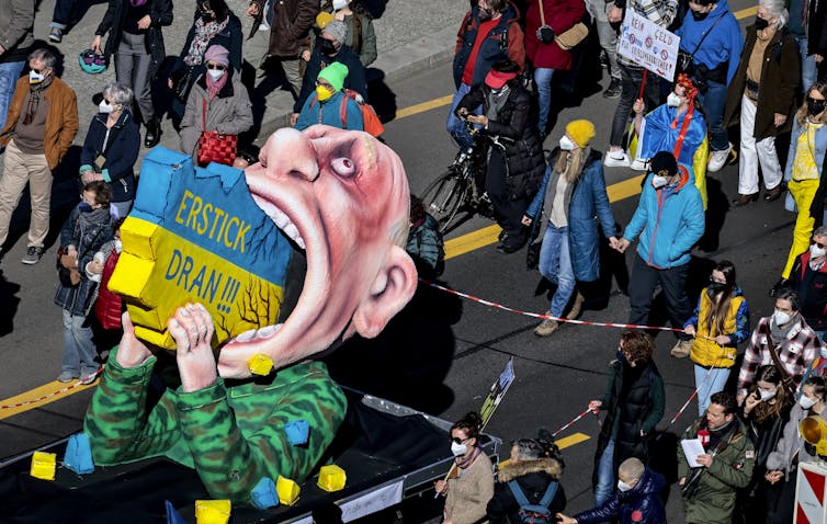 A large, caricaturish figure of Russian President Vladimir Putin swallowing a map of Ukraine is part of a demonstration, with people walking behind it.