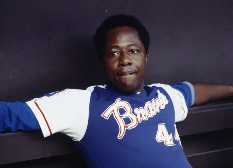 Man wearing blue baseball jersey sits in a dugout bench.