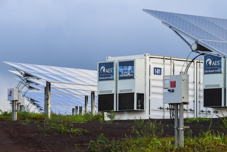 Rows of solar panels and two batteries the size of small shipping containers sit in a field.
