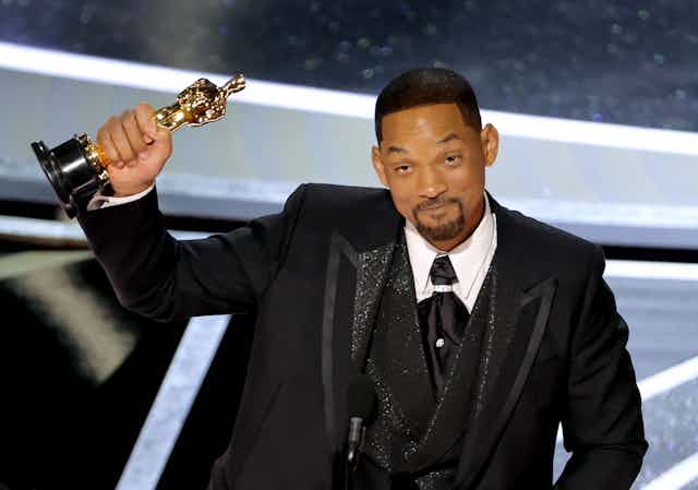 A black man dressed in a tuxedo is holding a small gold statue.