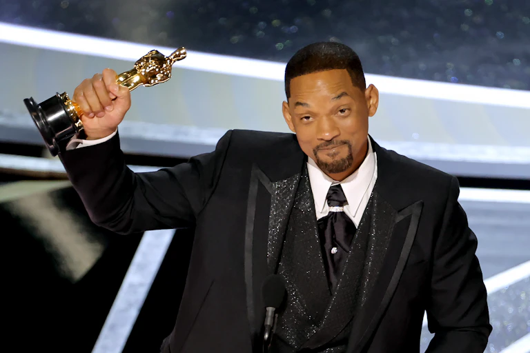 A black man dressed in a tuxedo is holding a small gold statue.