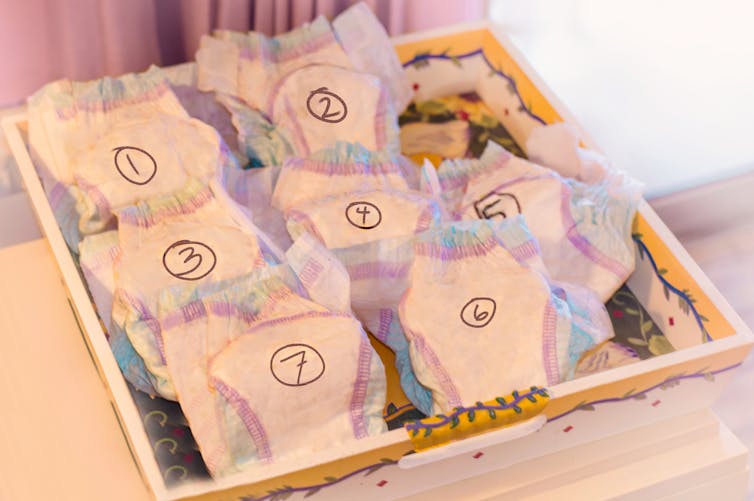 Seven disposable white baby diapers with colorful trim are grouped in a tray with numbers drawn on them in marker.