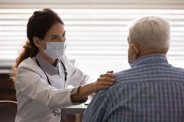 Una médico con mascarilla en consulta posa una mano en el hombro de un paciente mayor con pelo gris, camisa azul y mascarilla. 