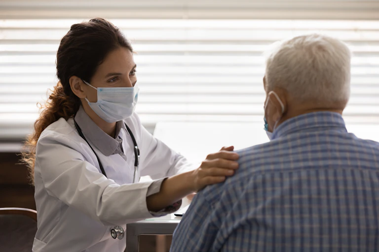 Una médico con mascarilla en consulta posa una mano en el hombro de un paciente mayor con pelo gris, camisa azul y mascarilla.