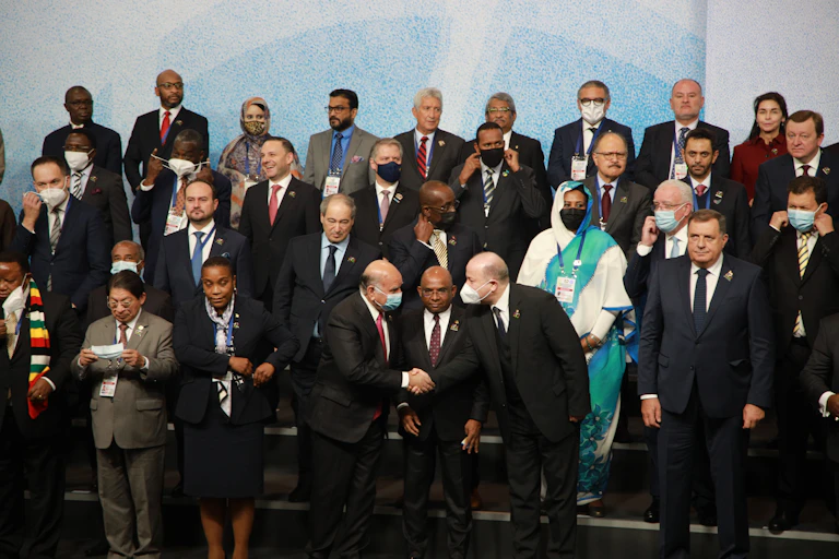 Leaders of non-aligned states pose for a photo, some shaking hands, most wearing masks.