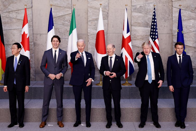 Six men in dark suits standing in front of six flags.