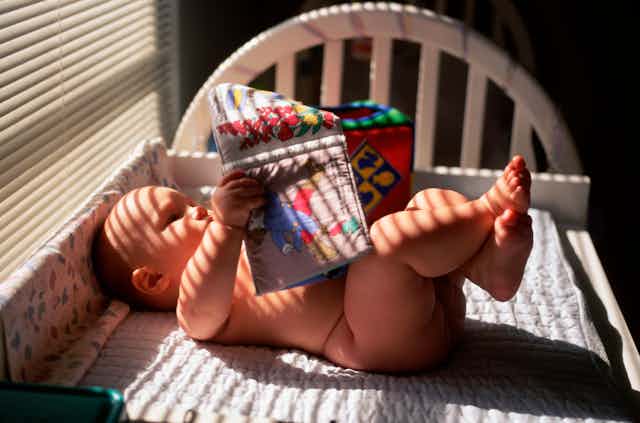 A baby lies naked on a changing table, engrossed in a cloth picture book, awaiting a diaper that may never come...
