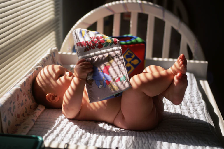 A baby lies naked on a changing table, engrossed in a cloth picture book, awaiting a diaper that may never come...