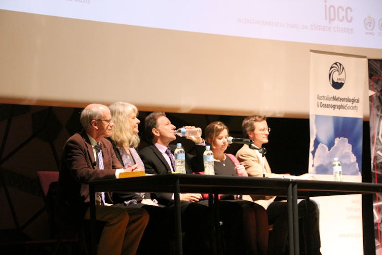 Five white people sit behind a table on a stage