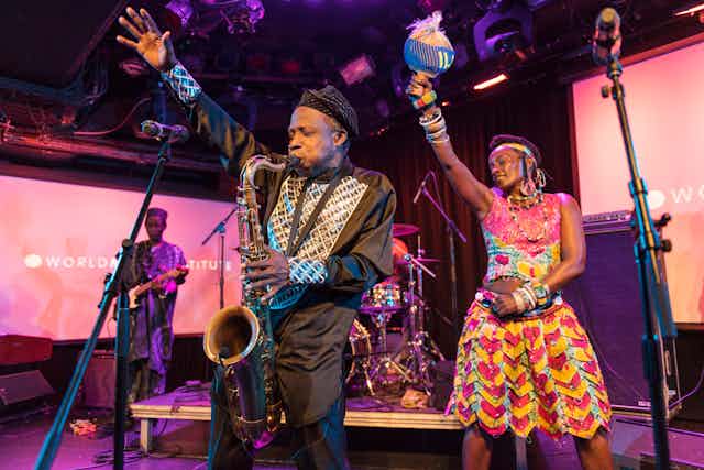 A band on stage, brightly coloured African attire. An elderly man blows into a saxophone. A woman next to him dances. Both raise their rights hands in the air.