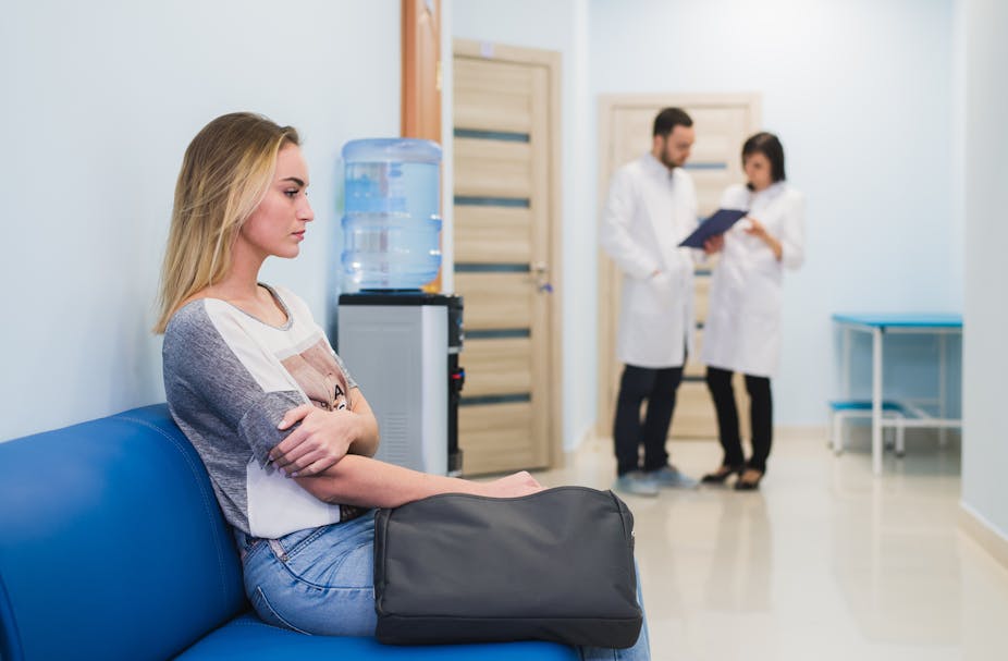Young woman sitting on a waiting room couch in the GP's office. Two GPs are in the background talking.