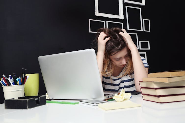 Woman holding her head in frustration while sitting at her desk, with her laptop, books and other office supplies set up in front of her.