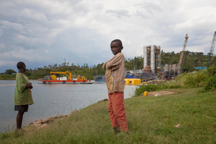 Two young boys stand on the grassy edge of a lake with an industrial structure in the background.