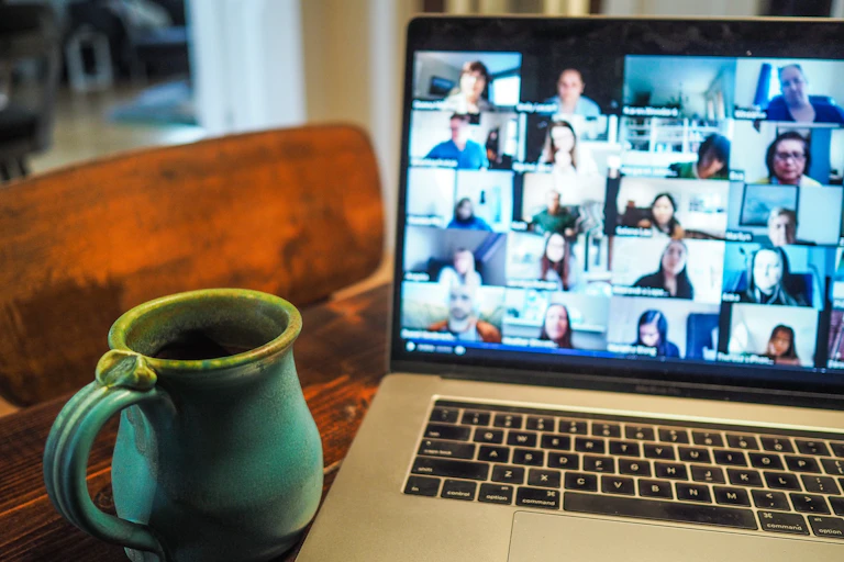 A coffee mug sits next to a laptop, the screen is displaying a zoom call with blurred faces.