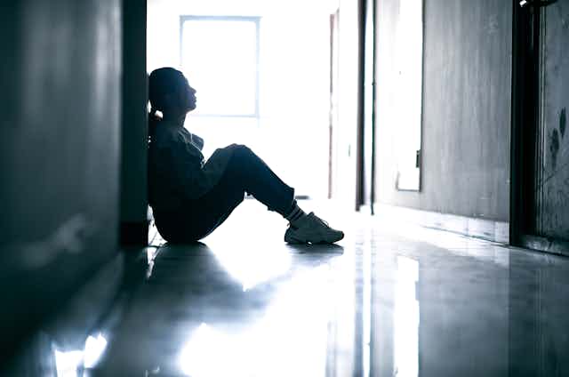 A sad teenage girl sits on the floor of a darkened hallway.