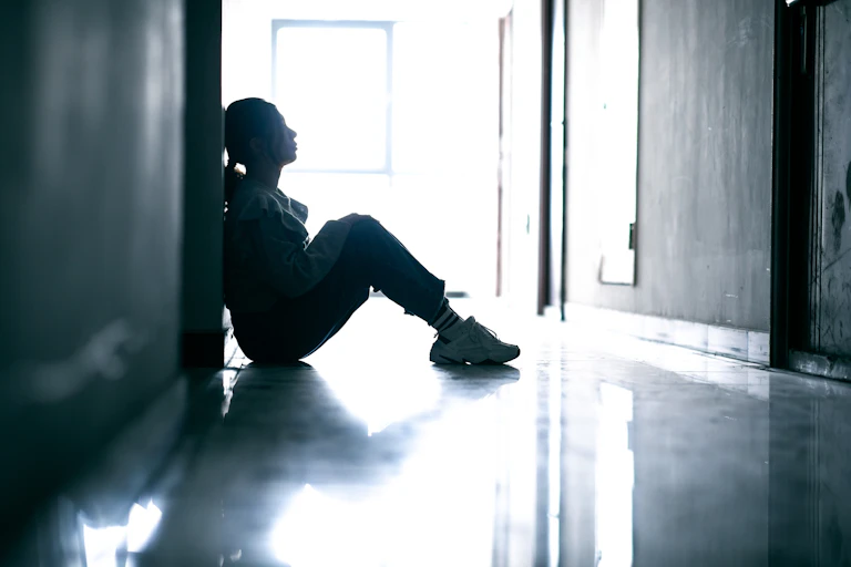 A sad teenage girl sits on the floor of a darkened hallway.