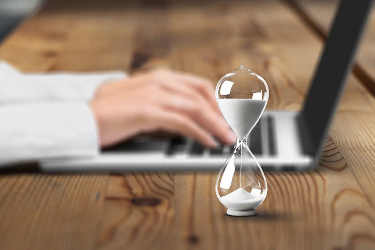an hourglass with white sand and a person's hands on laptop keyboard in the background