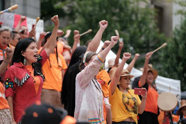 First Nations women in orange and red shirts protest outside, with fists raised.