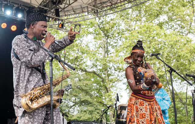 A man and a woman on stage in brightly patterned African attire. He sings into a microphone, a saxophone around his next. She holds a traditional musical instrument.