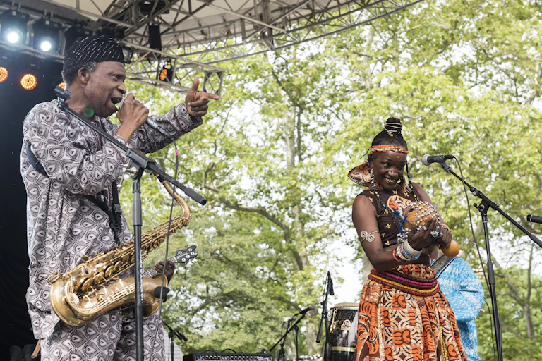 A man and a woman on stage in brightly patterned African attire. He sings into a microphone, a saxophone around his next. She holds a traditional musical instrument.
