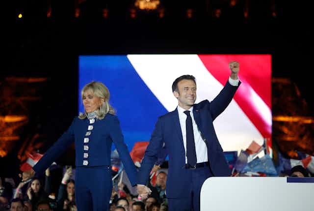 Emmanuel and Brigitte Macron waving to a crowd in front of a screen depicting a French flag.