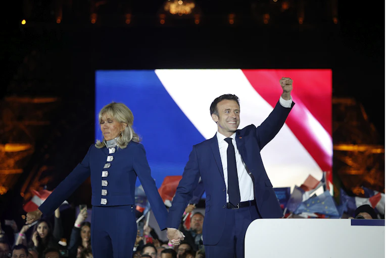 Emmanuel and Brigitte Macron waving to a crowd in front of a screen depicting a French flag.