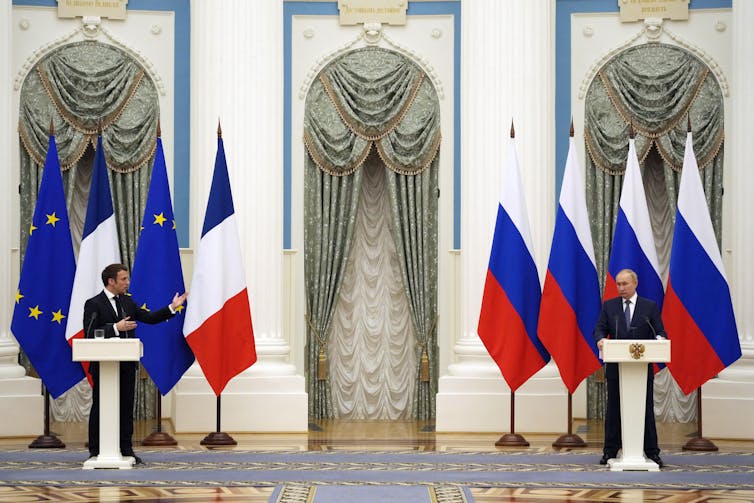 Emmanuel Macron and Vladimir Putin holding a joint press conference at two podiums placed at a great distance on a stage.