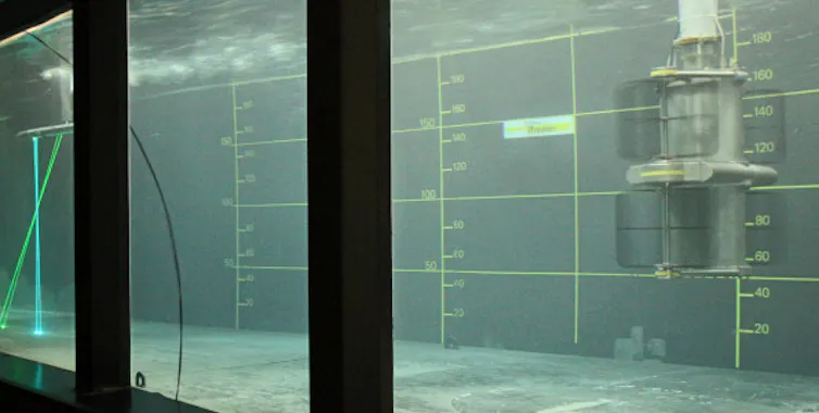 Vu d’un bassin rempli d’eau, dans lequel est immergé un élément métallique ; deux rayons laser sont visibles à une extrémité du bassin