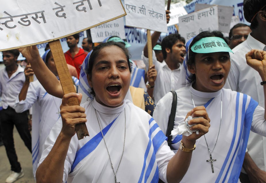 Two Christian nuns, dressed in white sarees with blue borders, holding placards, as they march ahead of people protesting with them.