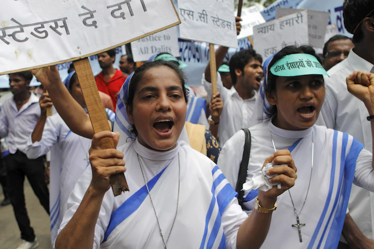 Two Christian nuns, dressed in white sarees with blue borders, holding placards, as they march ahead of people protesting with them.
