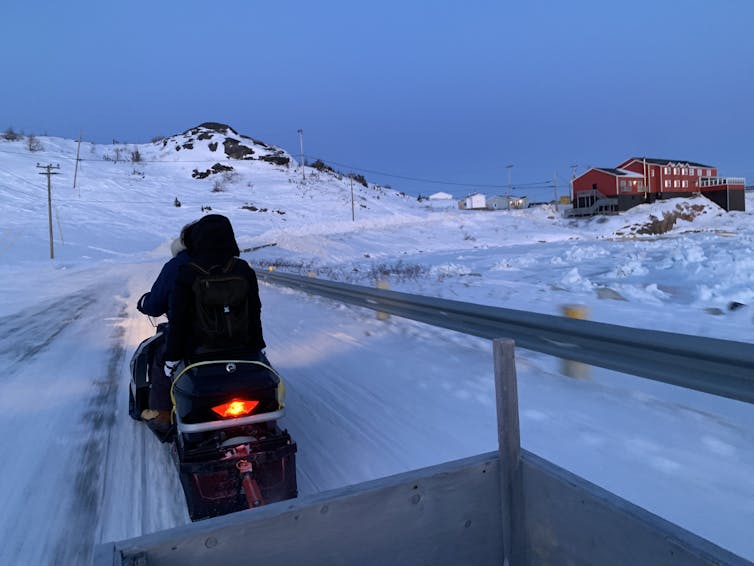 A person driving a snowmobile seen from behind, heading toward a red building in the distance.