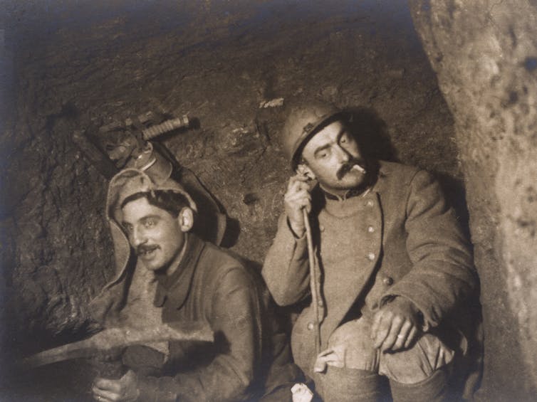 The black-and-white photo shows two World War I soldiers listening to a device while seated in a tunnel.