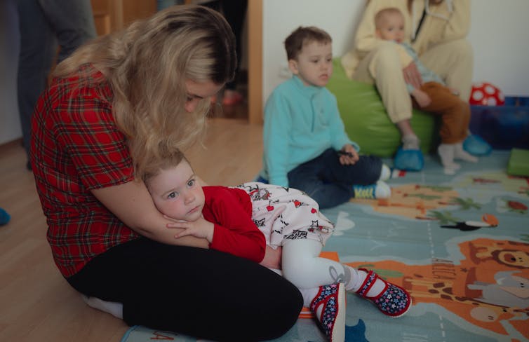 A blond woman holds a small child in her lap who lies against her mother's arms