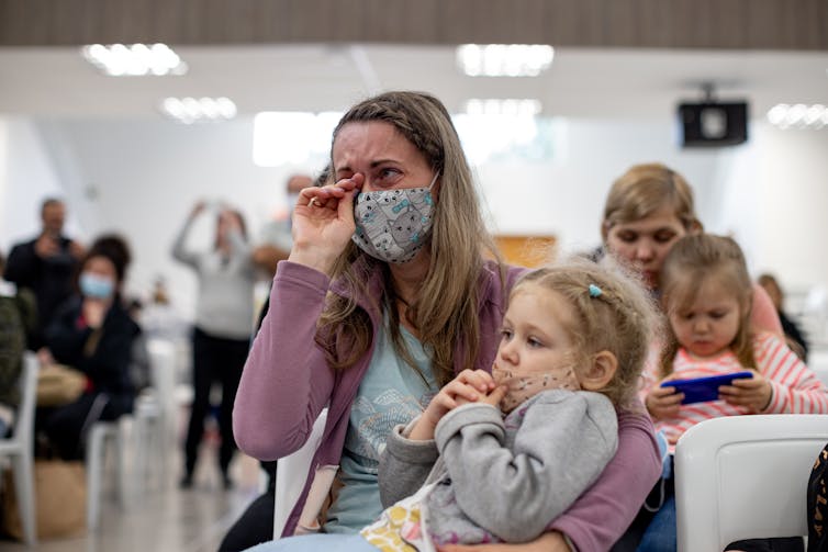 A blond woman wearing a face mask holds a child in her arms and wipes away tears, in a room of other people seated.