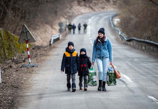 Two young boys and a woman carrying bags walk along an empty highway