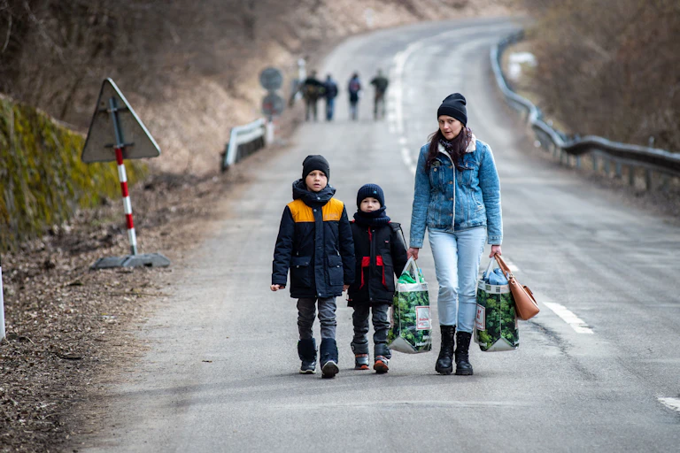 Two young boys and a woman carrying bags walk along an empty highway