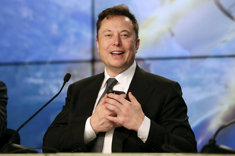 a man wearing a business suit stands smiling and pressing a smart phone to his chest with both hands