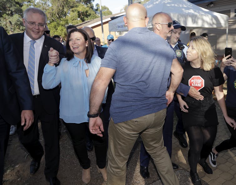 Morrison and his wife holds hands and smile on the left while a protester in a 'stop Adani' t-shirt is held back by security on the right.