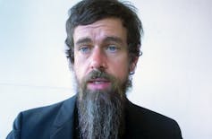 A white guy with a long beard and gray suit stares straight ahead while appearing to open his mouth to speak