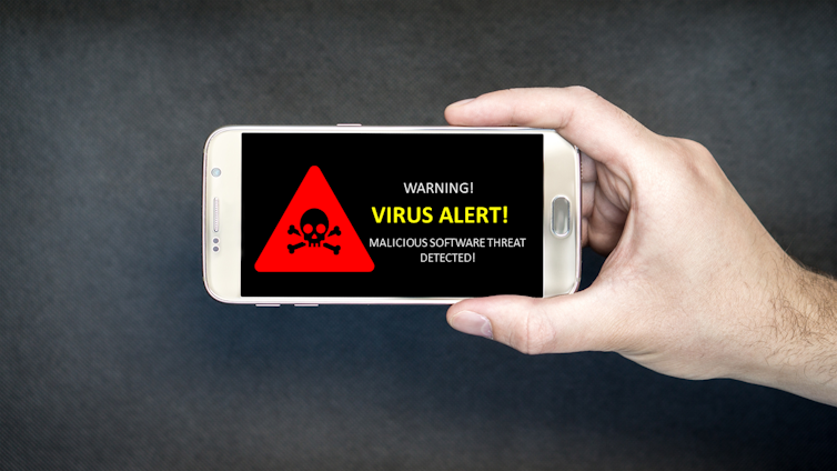 A smartphone with a virus alert warning is held up by a hand in front of a dark background.