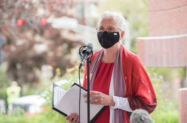 A woman wears a facemask and stands behind a microphone, she has white hair and is wearing a red top