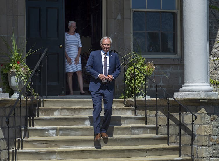 A man walks down steps, he's wearing a suit, there's a woman standing in the doorway behind him wearing a white dress.