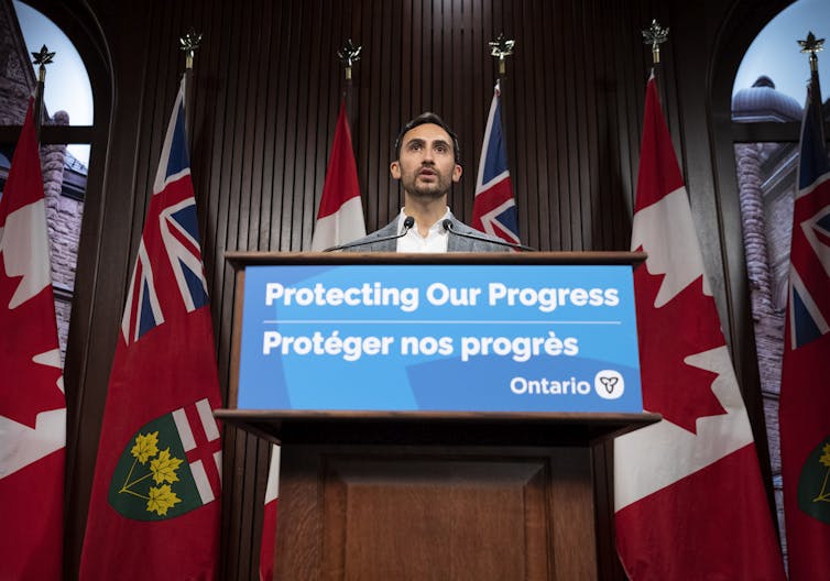 A man in a shirt and jacket is seen at a podium that says protecting our progress.