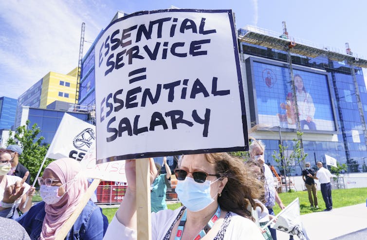 People in face masks holding signs. In the foreground, a woman holds a sign that says'Essential service = essential salary'