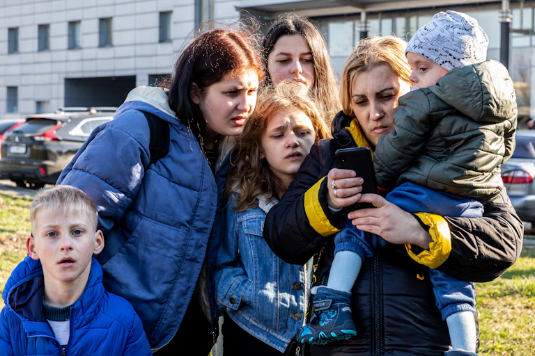 Two women and four children, outside in warm clothes, looking with concern at a mobile phone screen.
