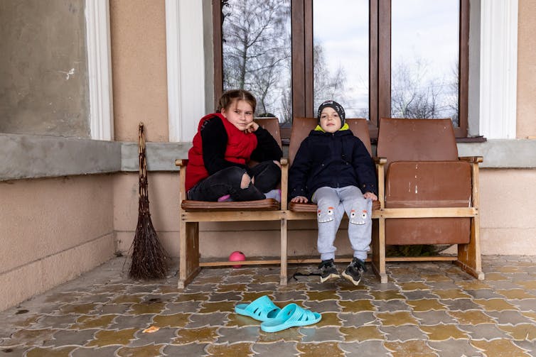 Two children sit on a bench on a tiled floor outside a building.