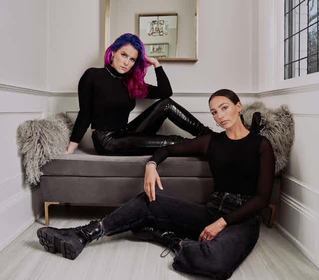 One woman with long purple hair sitting on a couch and one woman with tied back long brown hair and black combat boots sitting on the ground.
